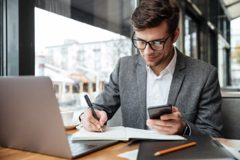 smiling businessman in eyeglasses sitting by the table in cafe with laptop computer while using smartphone and writing something